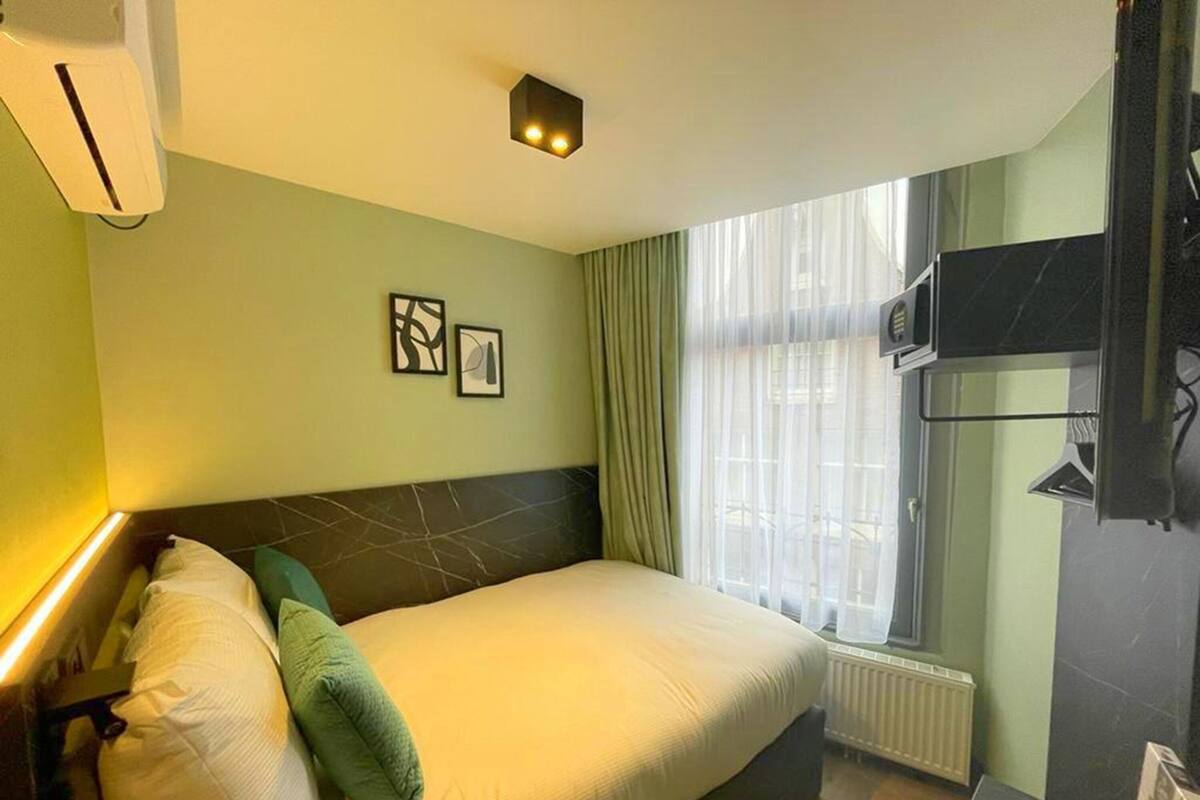 Habitación de hotel con una cama, un ventanal con cortinas translúcidas, un televisor de pantalla plana montado en la pared y un radiador debajo de la ventana.