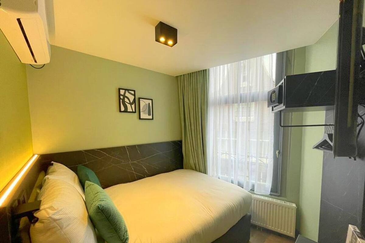 Habitación de hotel con cama, ventana con cortinas, radiador y televisor montado en la pared.