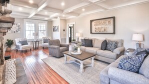 A spacious living room with a wooden floor, a grey sofa, a grey armchair, and a coffee table. There is a fireplace on the left and a large window in the background.