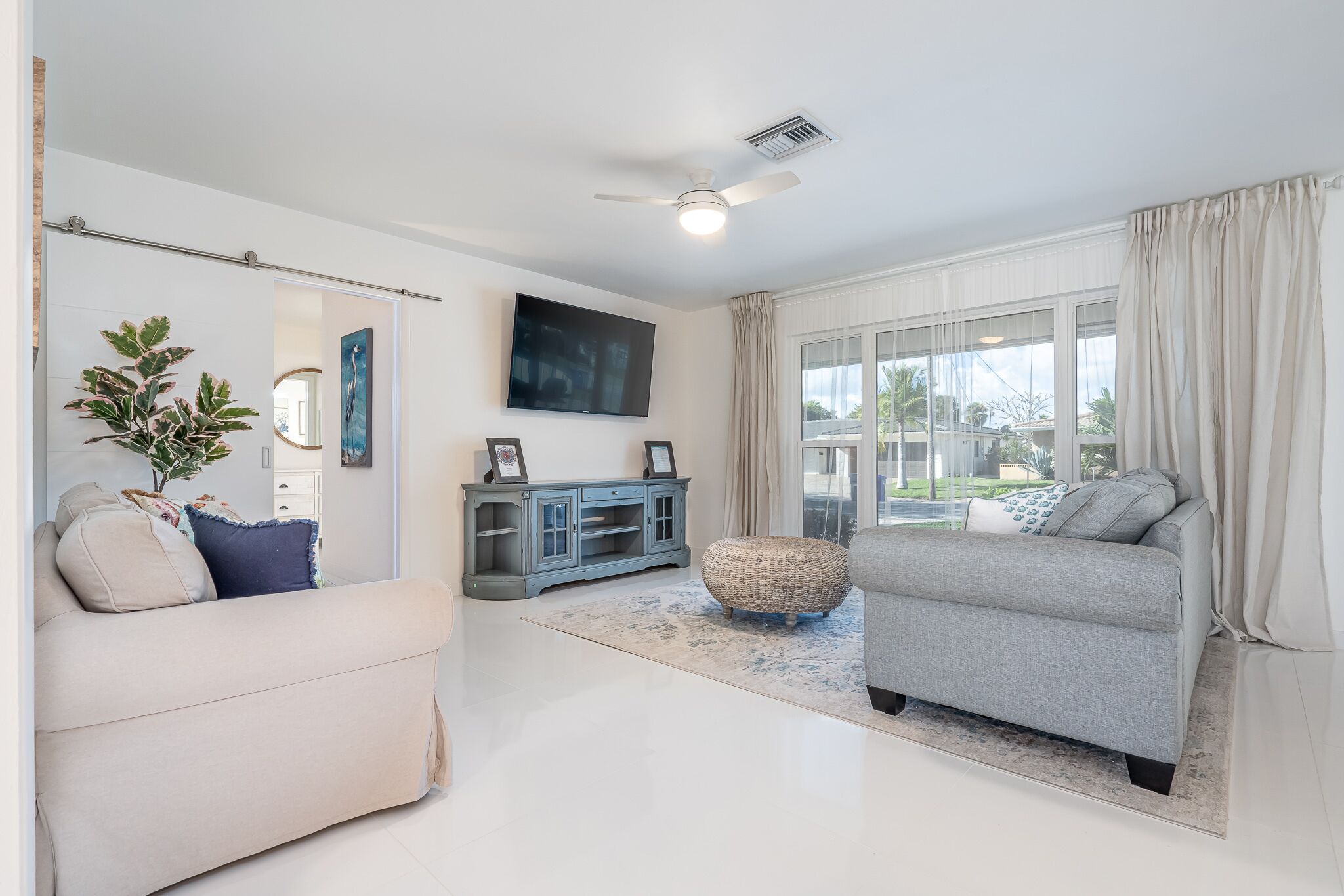 A modern living room with a grey sofa, a beige armchair, a flat-screen TV, a ceiling fan, and a large window with curtains.