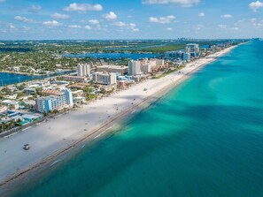 An aerial view of a coastal city with a long sandy beach, clear blue waters, and a mix of high-rise and low-rise buildings along the shoreline.