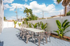 An outdoor patio with a long table and chairs, surrounded by plants and a white fence.