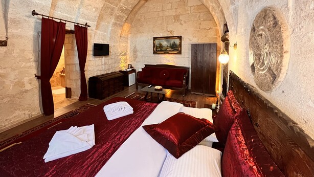 A room with a vaulted stone ceiling, a bed with red bedding, a wooden bench, a small table, and a painting on the wall. - Shadow Cave House (Ürgüp)
