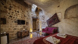 A stone-walled room with a bed, a sofa, a television, and a patterned rug. - Shadow Cave House (Ürgüp)