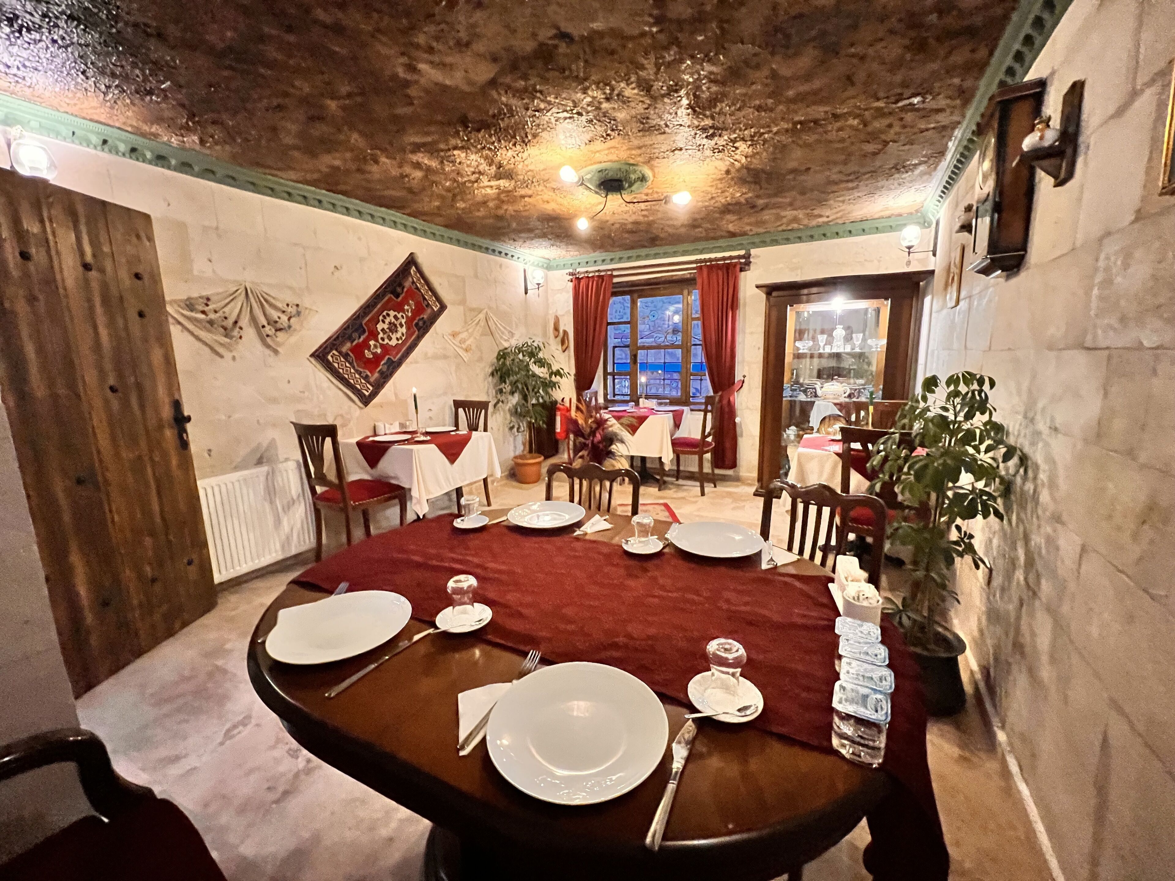 Uma sala de jantar com uma mesa de madeira posta para quatro pessoas, rodeada por cadeiras. Há plantas em vasos e uma janela com cortinas vermelhas.