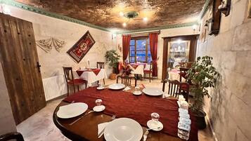 Uma sala de jantar com uma mesa de madeira posta para quatro pessoas, rodeada por cadeiras. Há plantas em vasos e uma janela com cortinas vermelhas.