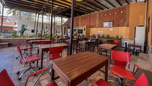 An outdoor cafe with wooden tables and red chairs, a brick wall with a TV, and a ceiling with exposed beams and lighting. - Hotel Oasis (Mazatlán)