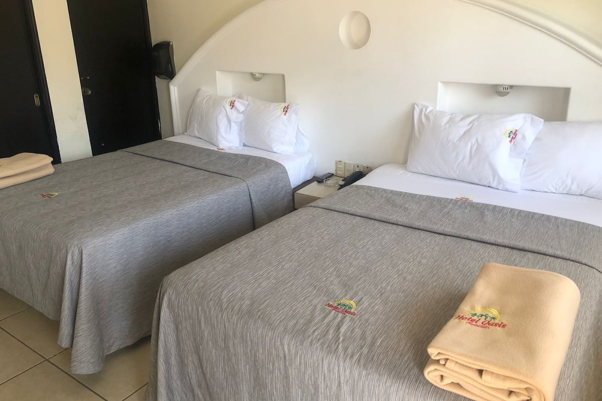 A hotel room with two beds, each with a grey bedspread and white pillows, a black door, and a white ceiling with a recessed light.