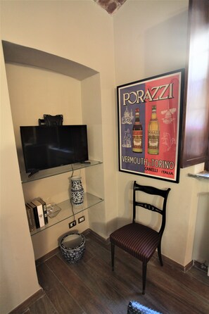 A room with a flat-screen TV on a glass shelf, a framed poster of a wine brand, a patterned vase, a wooden chair, and a small round table.