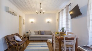 Una sala de estar con un sofá, una mesa de comedor con sillas, un sillón de mimbre, una araña y un televisor de pantalla plana montado en la pared.