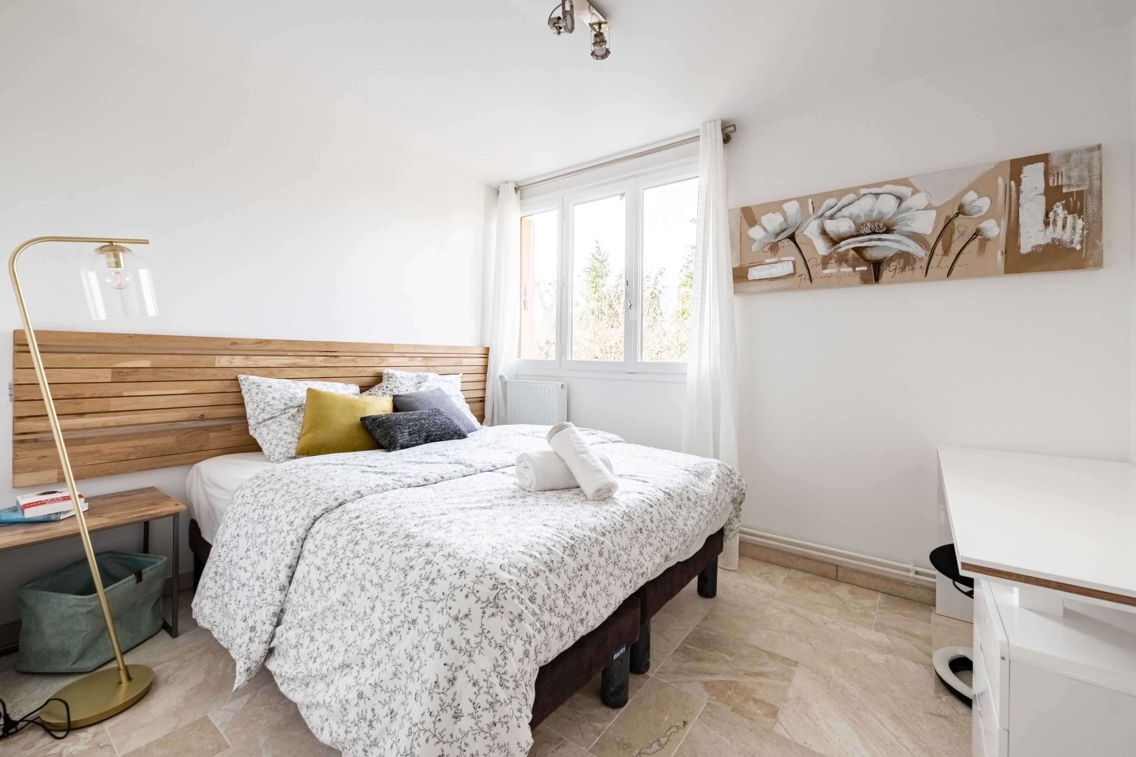 Une chambre à coucher avec une tête de lit en bois, un lit recouvert d’une literie à motifs floraux, une table de chevet avec des livres, une lampe sur pied et une fenêtre donnant sur un paysage.