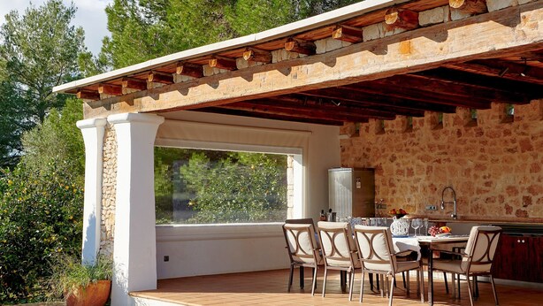 Un patio exterior con pared de piedra, pérgola de madera, una mesa con sillas y vistas a árboles.