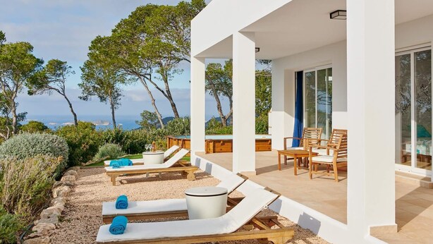 Un espace de détente extérieur moderne, doté de chaises longues et d’une piscine, offrant une vue panoramique sur un paysage verdoyant, avec des arbres et la mer à l’horizon.