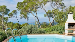 Un bain à remous en bois avec des échelles en métal, une table en verre avec une boisson et une serviette, et un barbecue, le tout entouré d’arbres et offrant une vue sur la mer.
