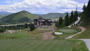 Un campo de golf con un edificio grande al fondo, rodeado de montañas y árboles.