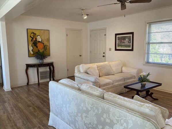 Ein Wohnzimmer mit einem beige farbenen Sofa, einem gemusterten Sofa, einem kleinen Tisch mit einer Vase, einem Bild an der Wand und einer Tür im Hintergrund.