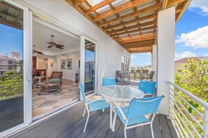Un patio techado al aire libre con un ventanal de vidrio que da a un comedor interior, amueblado con sillas azules y una mesa blanca.