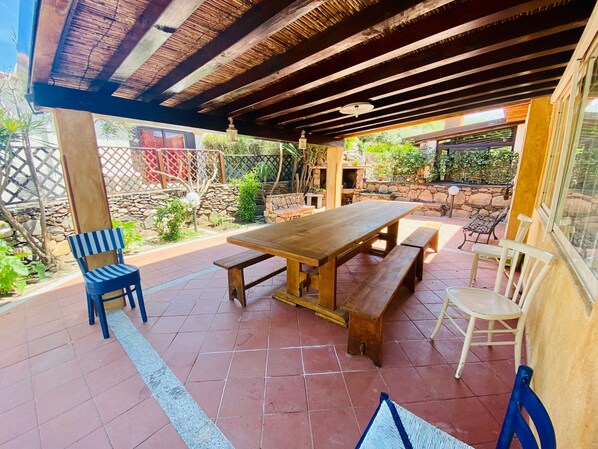 An outdoor patio with a wooden table and benches, surrounded by stone walls and potted plants.