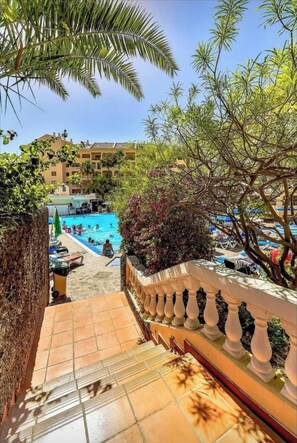 A pool area with a view of a building in the background, surrounded by trees and a tiled walkway.