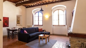 Una sala de estar con sofá negro, mesa de centro de madera y chimenea. Hay dos ventanas con arcos y un techo con vigas de madera a la vista.