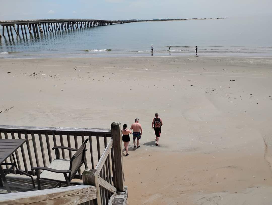 A beach with a wooden deck and chairs, a pier in the background, and a few people walking on the sand.