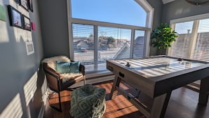 Ein gemütliches Zimmer mit Billardtisch, einem bequemen Sessel, einem großen Fenster mit Jalousien und einem Topf mit Pflanze.