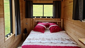 A wooden cabin bedroom with a bed, curtains, a window with a view of the outdoors, and a mounted deer head on the wall. - Cab'ânes du Pibeste (Segus)