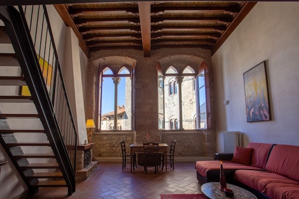 A spacious room with a wooden staircase, a dining area with a table and chairs, a red sofa, and large windows with a view of historical architecture.