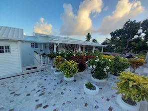 A white building with a covered patio area, surrounded by potted plants and a tiled floor.