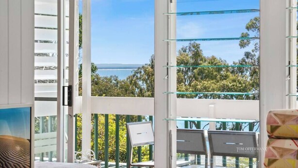 A balcony with a clear view of the sea and trees, a laptop on a table, and a chair.