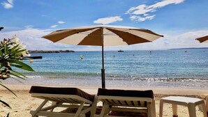 En strand med to liggestole og en stor parasol, med udsigt over havet med både og en klippeformation i baggrunden.