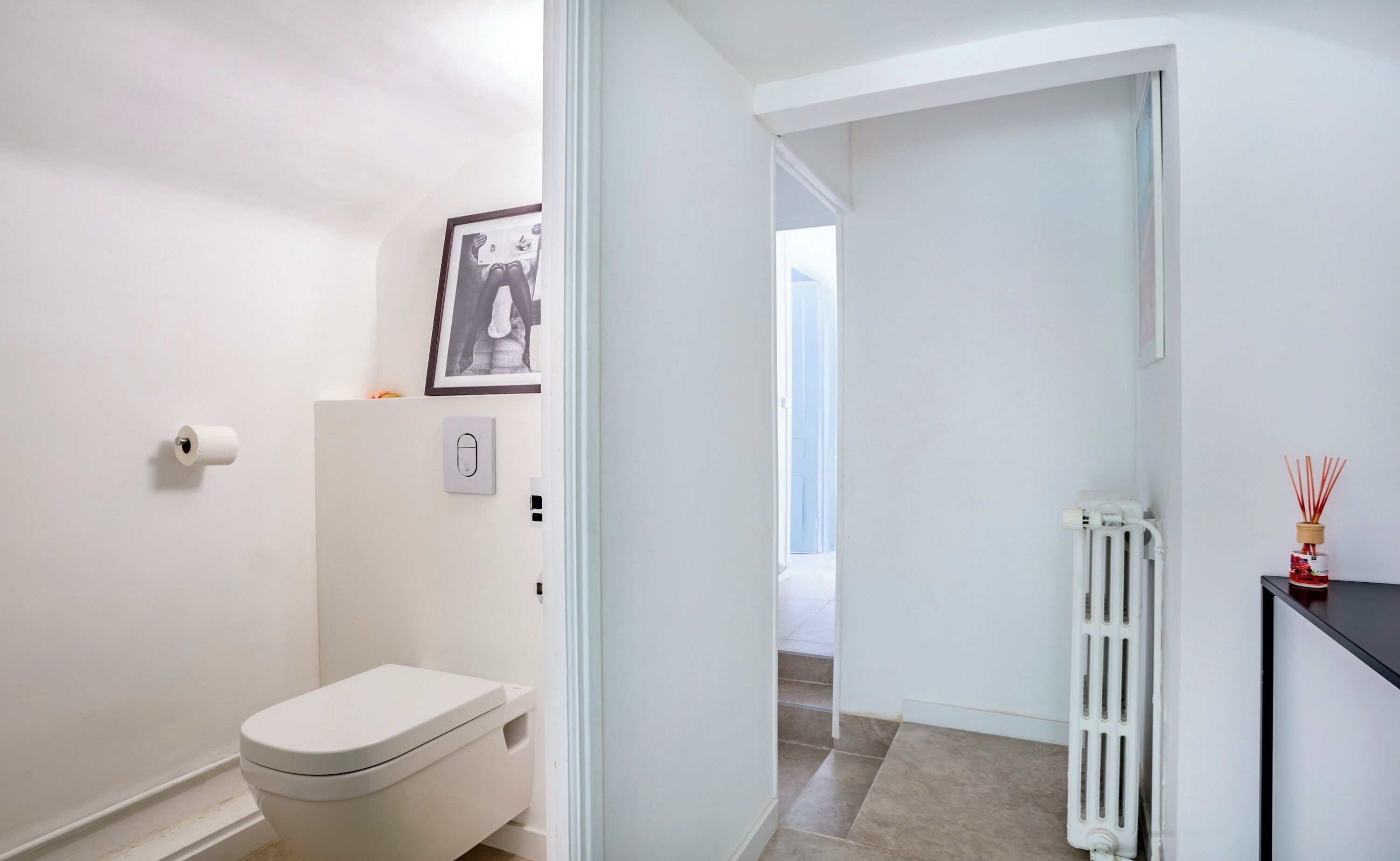 Un baño moderno con un inodoro, un cuadro enmarcado en la pared, un radiador y una pequeña mesa con una botella.