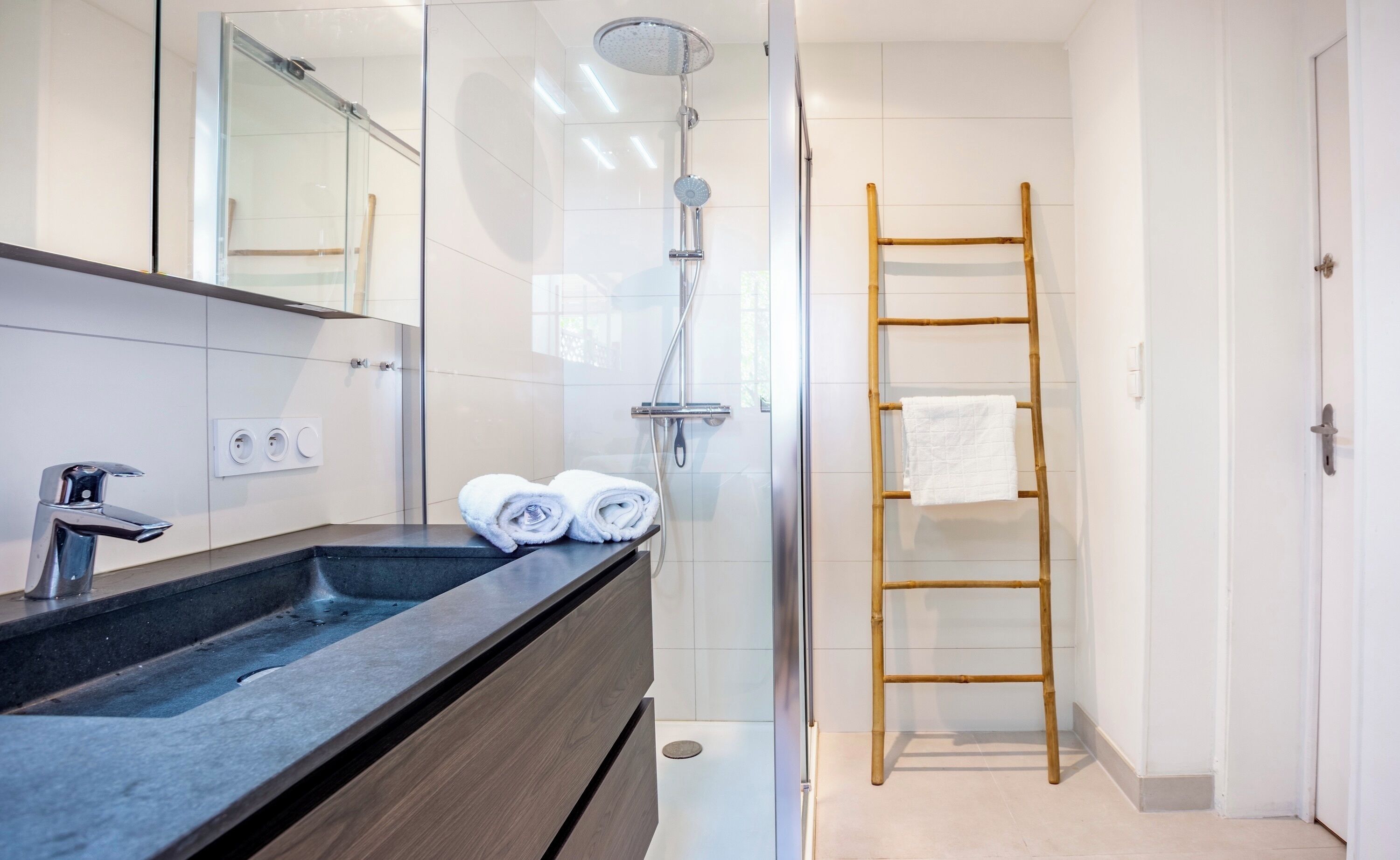 A modern bathroom with a glass shower enclosure, a wooden ladder, and a sink with two rolled towels.