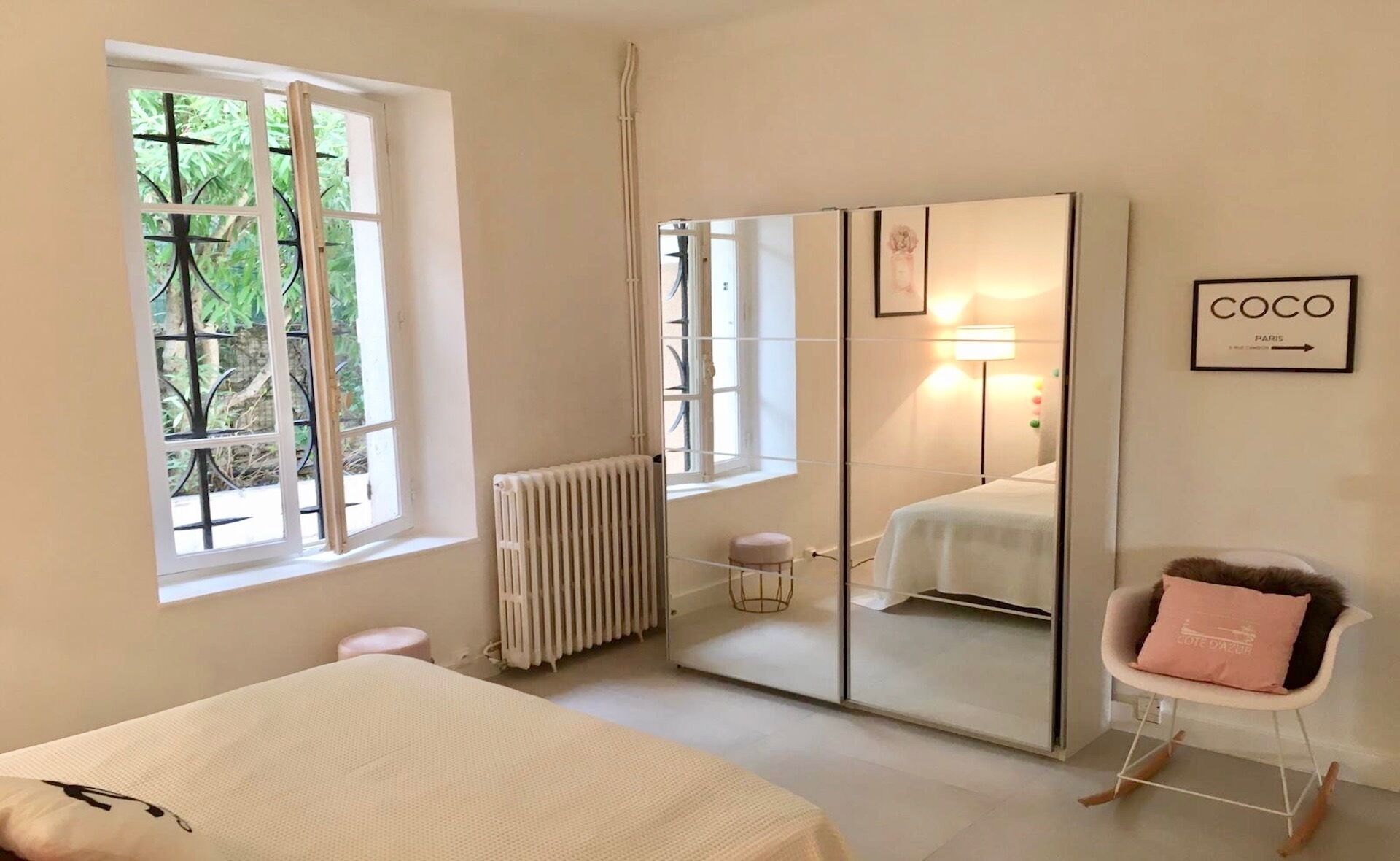 A bedroom with a bed, a wardrobe, a radiator, a window with a view of greenery, and a wall-mounted picture frame.