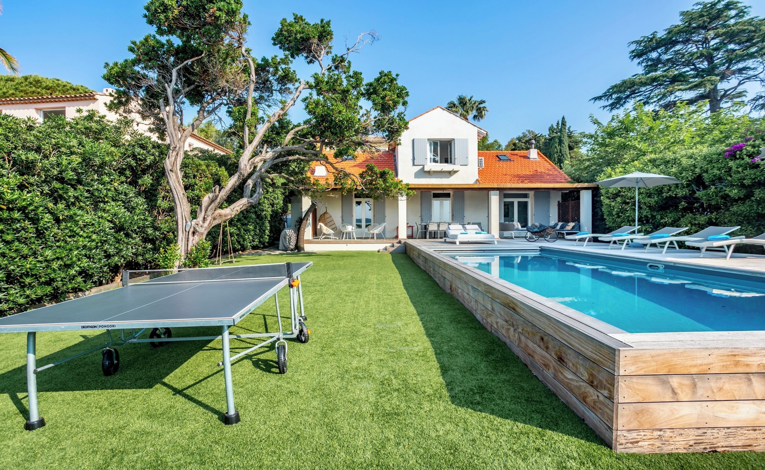 A pool area with a ping pong table, lounge chairs, and a house in the background.