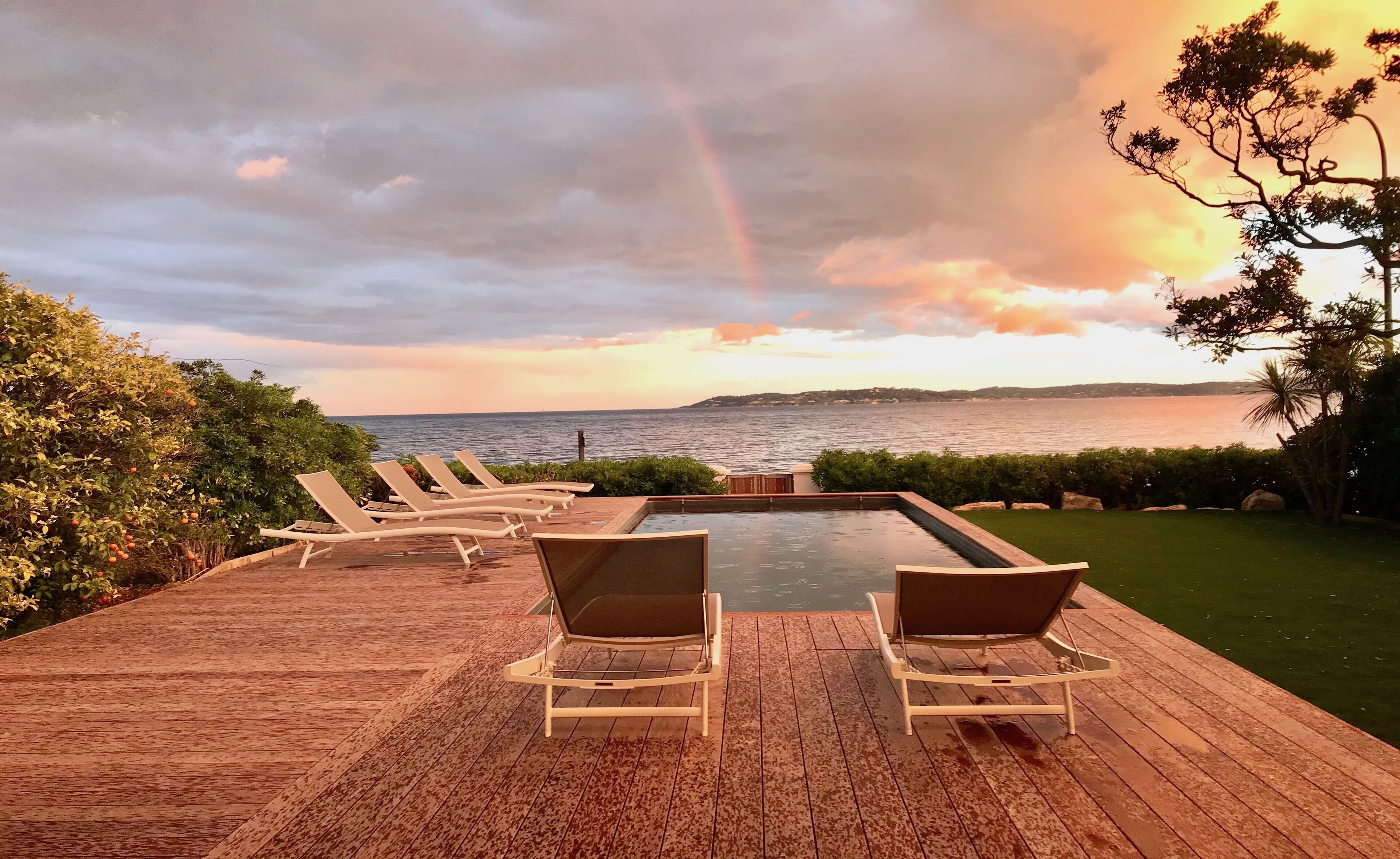 Une terrasse dotée de chaises longues donnant sur une piscine et la mer, avec un arc-en-ciel visible dans le ciel.