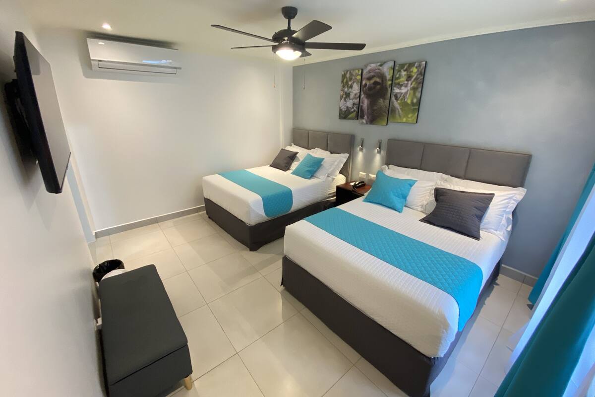 Habitación de hotel con dos camas, ventilador de techo, aire acondicionado y televisor de pantalla plana montado en la pared.