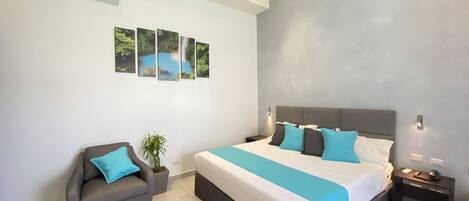 A modern bedroom with a large bed, a grey armchair, a small potted plant, and a wall-mounted air conditioner.