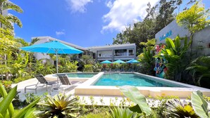 A pool area with lounge chairs and umbrellas, surrounded by lush greenery and a building with a sign. - Hotel Naoz (Quepos)