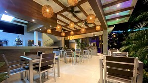 A modern restaurant with wooden ceiling beams, hanging wicker lamps, and a mix of wooden and metal furniture. - Hotel Naoz (Quepos)