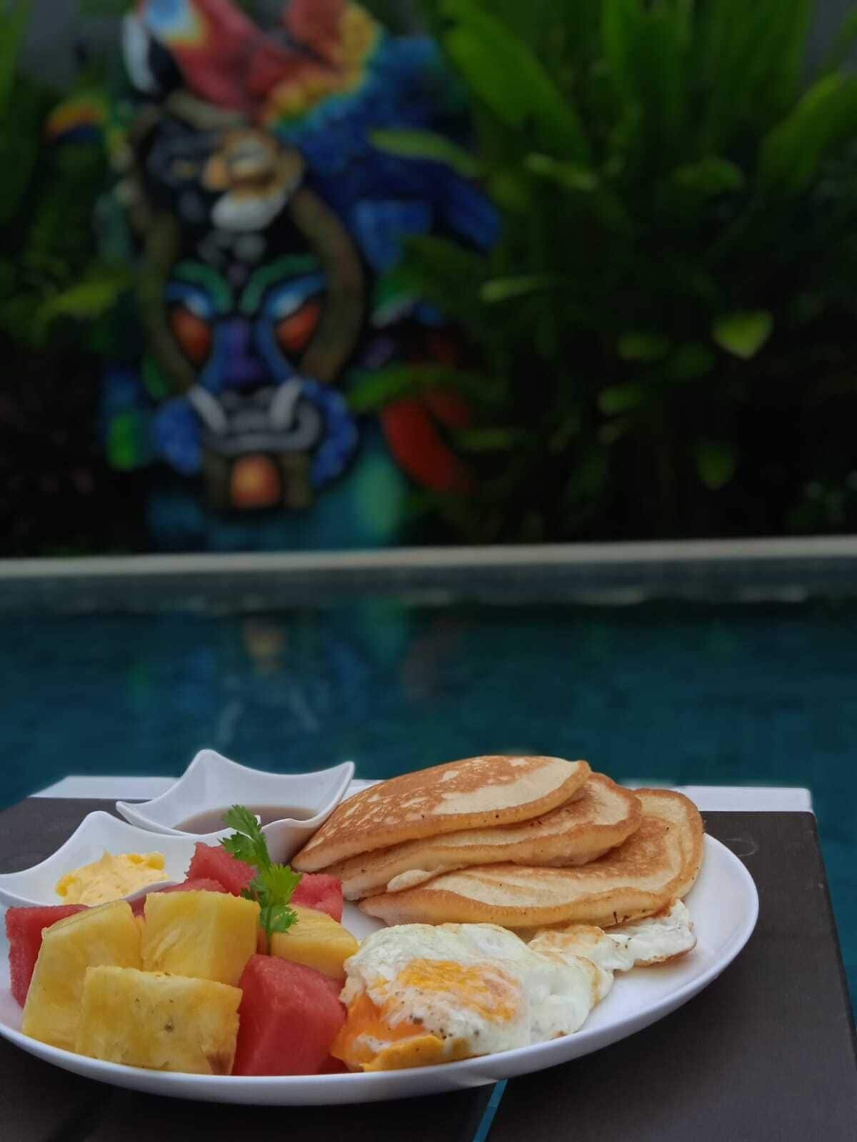 A plate with pancakes, eggs, and fruit, with a pool and greenery in the background.