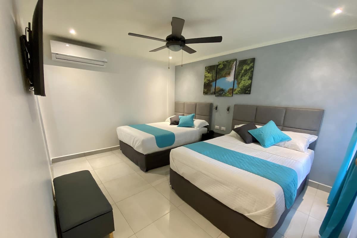 Habitación de hotel con dos camas, ventilador de techo, televisión de pantalla plana y una obra de arte en la pared.