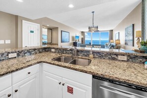 A kitchen with granite countertops, a double sink, and white cabinets. There is a view of the ocean through large windows.