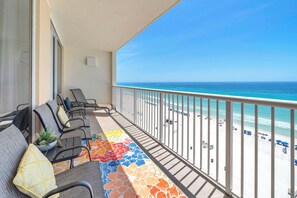 Un balcon avec des chaises et un tapis coloré, donnant sur une plage parsemée de parasols et fréquentée par des gens.