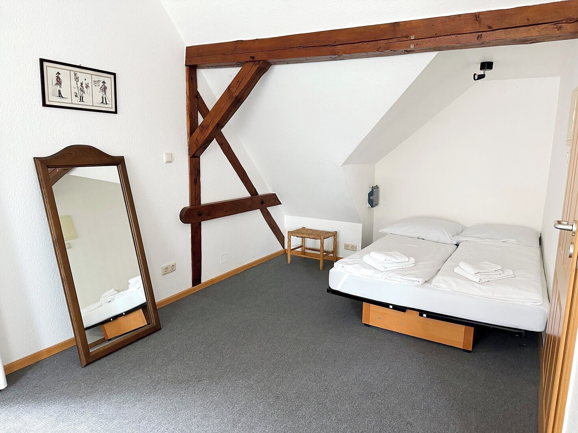 A bedroom with a bed, a wooden beam structure, a small table, a chair, a mirror, and a wall-mounted picture.