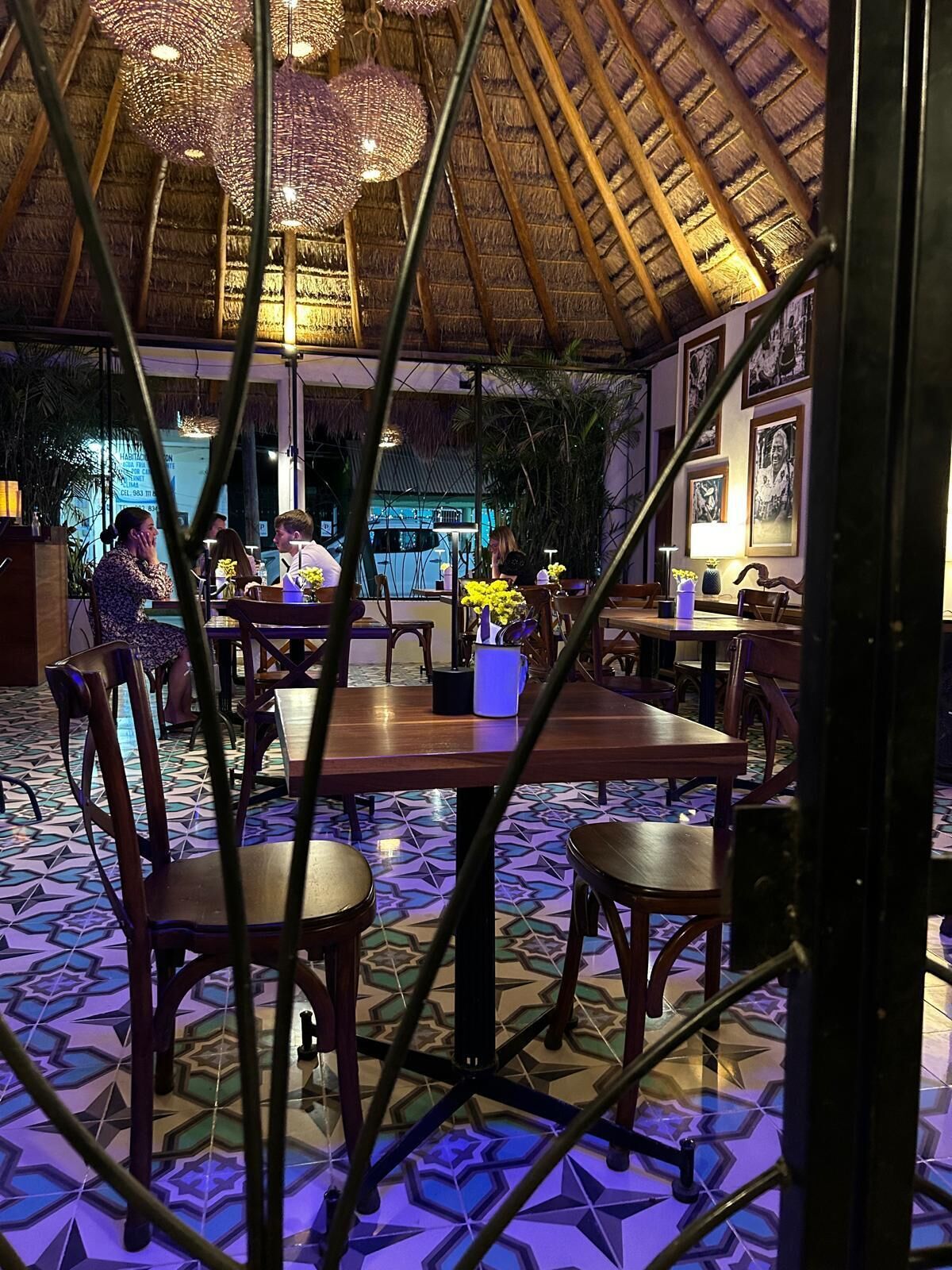 Un restaurante con techo de paja, mesas y sillas de madera, y un piso de baldosas con diseño geométrico.