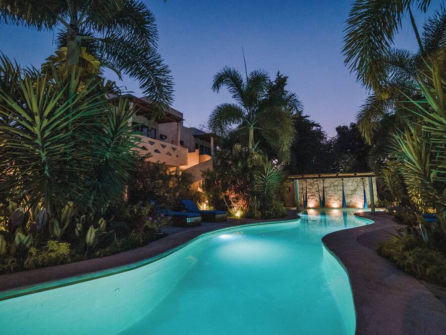 A swimming pool surrounded by tropical vegetation and a building with a stone facade.