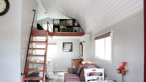 A loft-style bedroom with a loft bed, a wooden ladder, a sofa, a dining table with a vase of flowers, and a bookshelf. - Little House in the Pine Woods (Heavener)