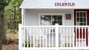 A white building with a red door labeled "IDLEWILD." There are white wicker chairs and a table on the porch. - Little House in the Pine Woods (Heavener)