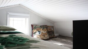 A bedroom with a sloped ceiling, a large window, and a bed with a floral bedspread. There are two decorative pillows on the bed. - Little House in the Pine Woods (Heavener)